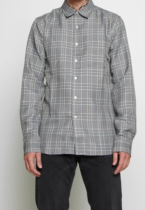 Chemise à manches longues boutonnée à carreaux gris et blancs avec une poche poitrine avant, portée avec un pantalon noir, tatouage visible sur le cou du porteur.