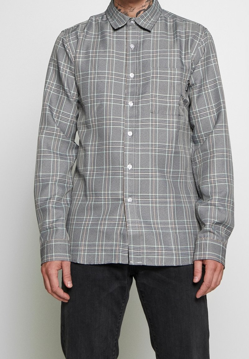 Chemise à manches longues boutonnée à carreaux gris et blancs avec une poche poitrine avant, portée avec un pantalon noir, tatouage visible sur le cou du porteur.