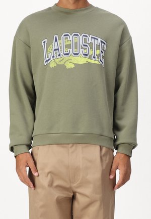 Homme portant un sweat-shirt vert olive avec un large texte "LACOSTE" et un logo crocodile jaune, assorti à un pantalon beige.