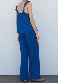 Femme portant un top sans manches bleu avec des bretelles volantes et un pantalon large assorti, se tenant sur un sol gris contre un mur blanc.