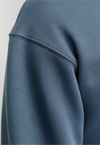 Gros plan d'une manche de sweat-shirt bleu montrant un tissu doux et texturé ainsi qu'une couture visible près de l'épaule.