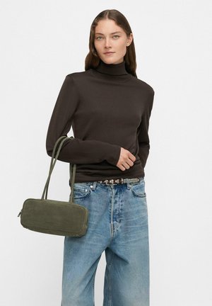 Frau in schwarzem Rollkragenpullover und blauen Jeans hält eine rechteckige, olivgrüne Handtasche mit zwei Griffen vor einem einfarbigen Hintergrund.