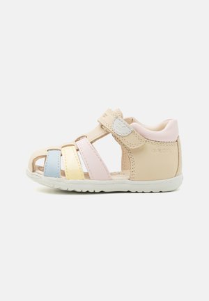 Sandálias de meninas em pele bege com tiras multicoloridas (azul, amarelo, rosa) e fecho de velcro, apresentando uma sola de borracha branca.