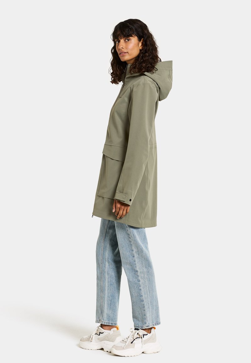 Grüne wasserdichte Jacke mit Kapuze, Fronttaschen und lockerer Passform, kombiniert mit hellblauen Jeans und weißen Sneakers.
