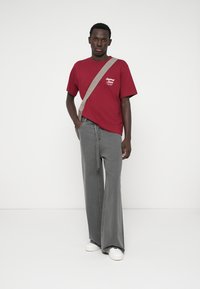 Bordo kokvilnas T-krekls ar baltu tekstu, apvienots ar pelēkiem platiem džinsu biksēm un baltām sporta apaviem. Modelim ir mugurā bezkrāsaina plecu soma.