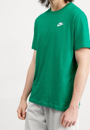 Personne portant un t-shirt Nike vert et un pantalon gris clair, debout les bras détendus devant un fond blanc.