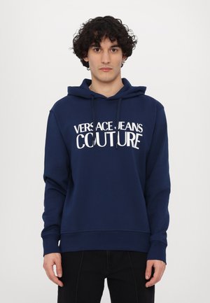 Mørkeblå hettegenser laget av mykt stoff, med en frontlomme i kengurustil, snorhette og stor hvit tekst "VERSACE JEANS COUTURE."
