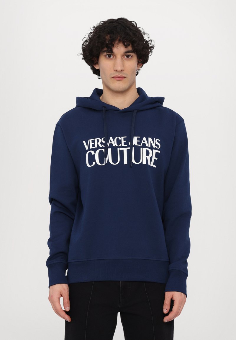 Mørkeblå hettegenser laget av mykt stoff, med en frontlomme i kengurustil, snorhette og stor hvit tekst "VERSACE JEANS COUTURE."