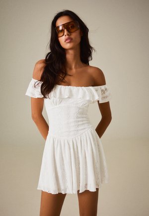 Femme portant une robe blanche en dentelle épaules dénudées et de grandes lunettes de soleil marron, debout devant un fond uni.