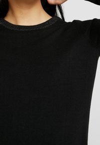 Zwarte trui met een geribbelde textuur en een glanzend zwart accent bij de kraag. De stof lijkt zacht en aansluitend.