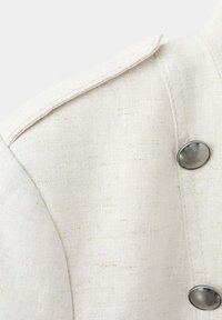 Gros plan d'une épaule de veste en tissu texturé beige clair avec un rabat et deux boutons métalliques ronds.
