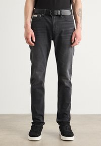 Schwarze Skinny-Jeans mit einem dezenten Waschung, kombiniert mit einem schwarzen Gürtel und schwarzen Wildledersneakern mit einer weißen Sohle, stehen vor einem neutralen Hintergrund.