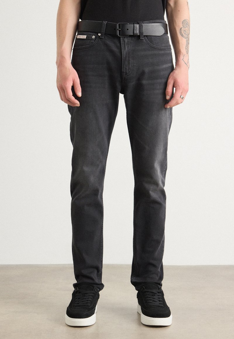 Schwarze Skinny-Jeans mit einem dezenten Waschung, kombiniert mit einem schwarzen Gürtel und schwarzen Wildledersneakern mit einer weißen Sohle, stehen vor einem neutralen Hintergrund.