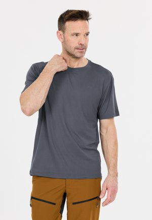 Man draagt een grijs shirt met korte mouwen en een bruine broek, raakt zijn nek aan met zijn linkerhand en kijkt naar rechts tegen een effen witte achtergrond.