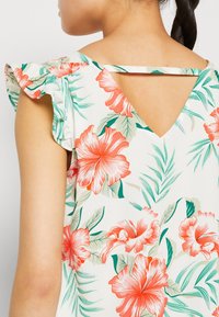 Blumenbluse mit Rüschensäumen, aus einem weißen Stoff mit großen orangefarbenen Hibiskusblüten und grünen Blättern. V-förmiger Rückendetail.