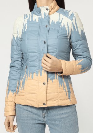 Mujer con chaqueta acolchada azul y beige con patrón de goteo de pintura abstracta, botones en el frente, bolsillos con cremallera y jeans de color azul claro.