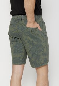Žalios šortai, pagaminti iš audinio su palmės lapų raštu, su šoninėmis kišenėmis ir elastinga juosmens juosta patogumui.