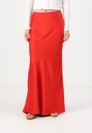 Kobieta w długiej, jaskrawoczerwonej spódnicy, białym krótko skróconym swetrze oraz beżowych sandałach, stojąca na tle jednolitej białej ściany.