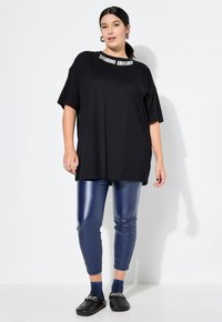 Zwarte oversized t-shirt met "STUDIO UNITED" in het wit bij de kraag, gecombineerd met glanzende marineblauwe leggings en zwarte instap schoenen.