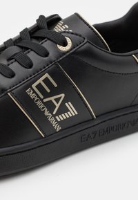 Black leather sneaker with gold accents, featuring a logo on the side, black laces, and a textured rubber sole. Simple and modern design.