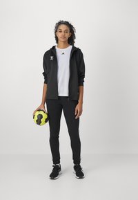 Kvinde i sort træningsdragt og hvid skjorte holder en gul og sort fodbold og står mod en ensfarvet hvid baggrund.