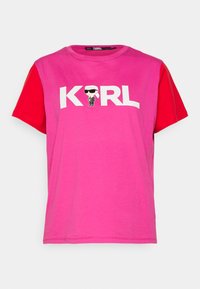 Růžové a červené tričko s krátkým rukávem s velkými bílými písmeny píšícími "KARL" a karikaturou postavy v brýlích. Bavlněný materiál.