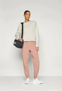 Žena v béžovej mikine, prašných ružových teplákoch, bielych teniskách, nesúca čiernu ramennú tašku Adidas, stojí na pozadí bez vzoru.