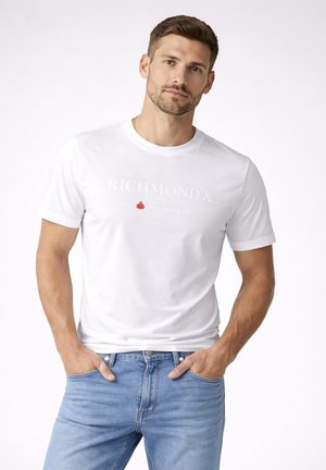 Uomo con indosso una t-shirt bianca Richmond X e jeans azzurri chiari, in piedi con le mani nelle tasche, espressione neutra su uno sfondo bianco semplice.