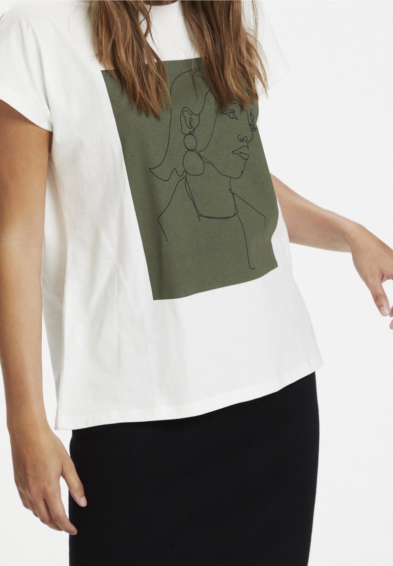 Femme porte un t-shirt blanc ample avec un carré vert olive présentant un dessin au trait d'une femme avec de grandes boucles d'oreilles, associé à une jupe noire.