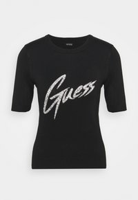 Černé krátké rukávy pletené tričko s kulatým výstřihem, s velkým stříbrným glitrovým logem "Guess" přes hrudník.