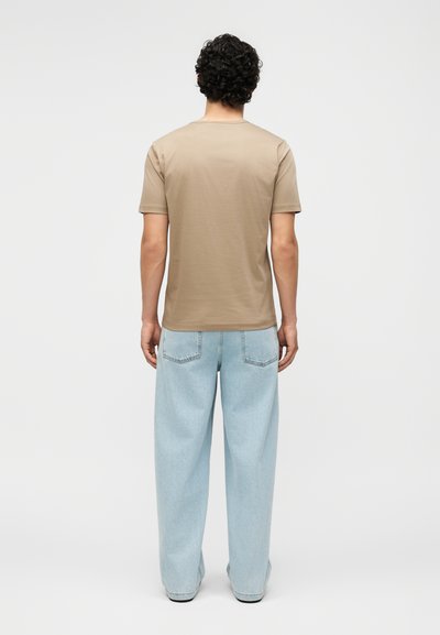 Άνδρας με σγουρά μαλλιά που φοράει απλό μπεζ κοντομάνικο μπλουζάκι και ανοιχτό μπλε τζιν με φαρδιά μπατζάκια, στέκεται γυρισμένος προς τα πίσω έναντι λευκού φόντου.