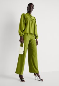 Blusa verde lime con maniche a sbuffo e scollatura con laccetto, abbinata a pantaloni larghi dello stesso colore. La modella tiene in mano una piccola borsa bianca di perline e indossa tacchi argentati.