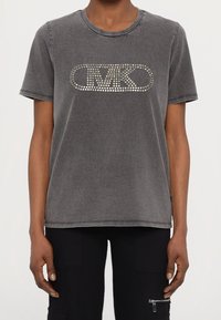 Camiseta de manga corta gris de tejido suave, con un gran diseño de logo metálico con tachuelas en el pecho y cuello redondo acanalado.