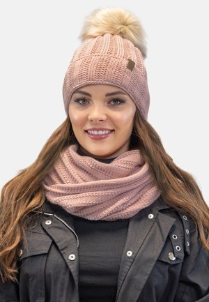 Bonnet tricoté rose clair avec un pompon duveteux et une écharpe assortie, présentant un motif texturé, porté sur une veste noire.