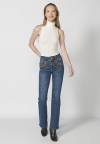 White sleeveless turtleneck top paired with dark blue flared jeans featuring embellished pocket details. Model stands in black heeled boots.