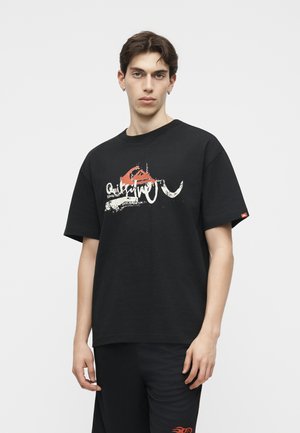 Νέος άνδρας που φοράει μαύρο t-shirt Quiksilver με γραφικά και μαύρο σορτς με κόκκινη φλόγα, στέκεται μπροστά σε λευκό φόντο.