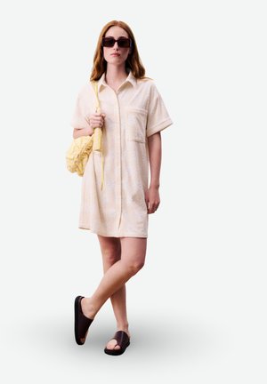 Femme portant une robe chemise crème à manches courtes, des sandales marron à enfiler, des lunettes de soleil, et tenant un sac à bandoulière jaune, debout avec les jambes croisées.