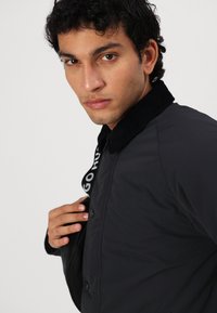 Casaco preto de nylon com colarinho de veludo cotelê, apresentando um fecho dianteiro com botões e uma alça com marca. Textura suave com um ajuste estruturado.