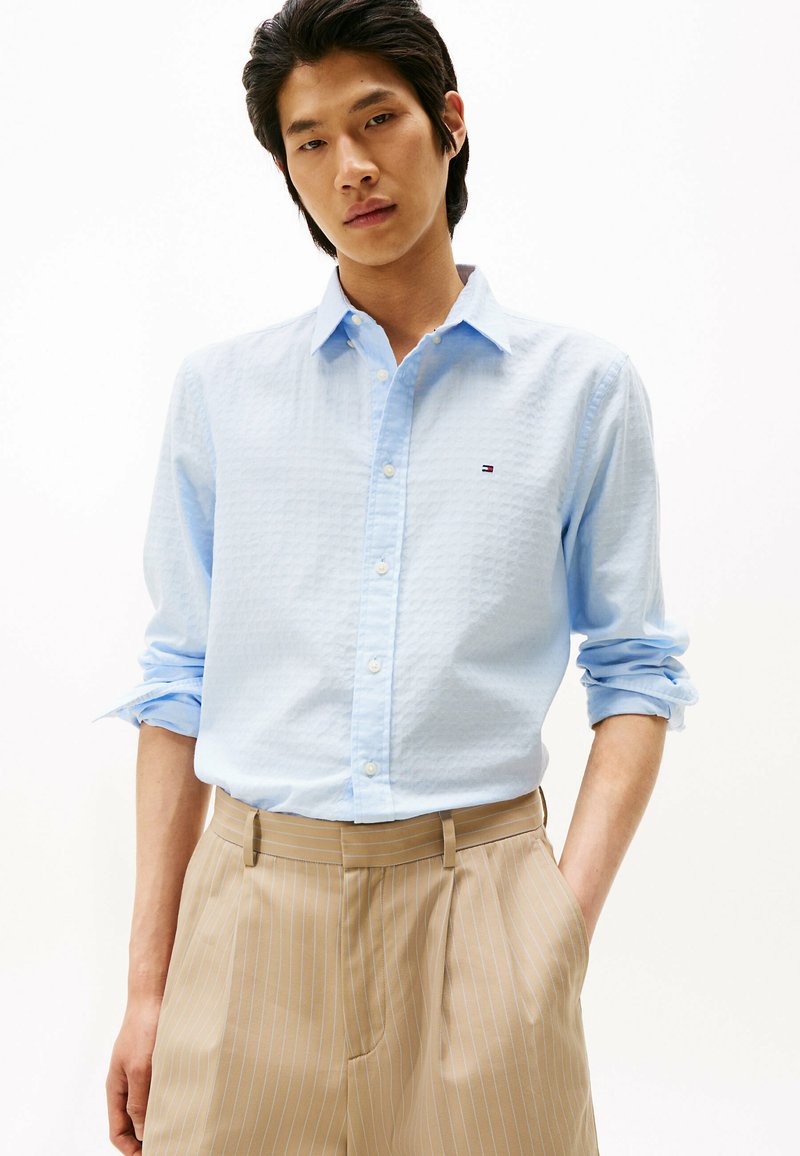 Camisa de botones azul claro con un patrón texturizado y un pequeño logo en el pecho, combinada con pantalones beige a rayas.