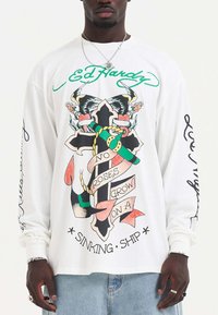 Weißes Langarm-T-Shirt mit einem zentralen Motiv aus einem Kreuz, Schlangen und Rosen, begleitet von Text. Akzentuiert mit grünen und roten Details.