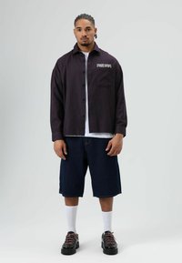 Zwart-rood geruit overhemd met lange mouwen, knoopsluiting aan de voorkant en geborduurd "FREEDOM" op de borst, gecombineerd met donkere denim shorts en zwarte schoenen.