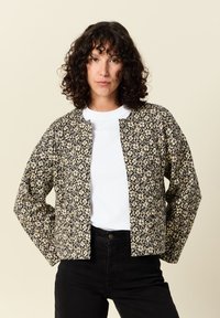 Czarna i złota kurtka w kwiatowy wzór o luźnym fasonie, z okrągłym kołnierzem i otwartym przodem, noszona na białej koszuli i czarnych spodniach.