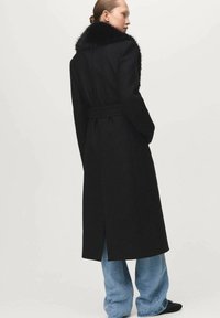 Lange zwarte jas met een bontkraag, een ceintuur in de taille en een split aan de achterkant, gecombineerd met losse blauwe jeans. Zachte textuur en strak ontwerp.