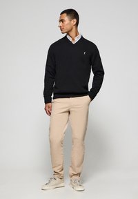 Suéter negro de cuello en V en un tejido suave, combinado con pantalones de algodón beige claro y zapatillas de color crema. Detalle sutil del logo en el suéter.