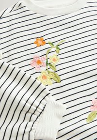 Pull blanc avec fines rayures horizontales noires et fleurs brodées rose, jaune et orange avec des feuilles vertes près de la poitrine et du poignet de la manche.