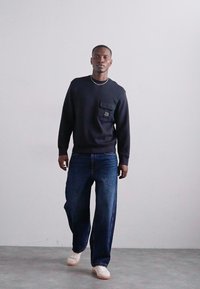 Jūros mėlynos spalvos apsiaustas su apvalia iškirpte ir priekiu su kišene, derinamas su plačiomis tamsiai mėlynomis džinsinėmis kelnėmis ir baltomis sportinėmis bateliais, stovintis prieš neutralų foną.