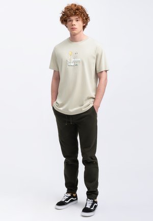 T-shirt beige clair avec un imprimé graphique, associé à un jogging vert foncé et des baskets noires avec des accents blancs et jaunes.