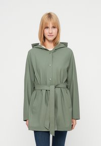 Νεαρή γυναίκα που φοράει αδιάβροχο με ζώνη σε απόχρωση φασκόμηλου και κουκούλα, στέκεται μπροστά σε ένα απλό λευκό φόντο.