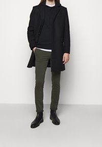 Schwarzer Überzieher über einem dunklen Hoodie, kombiniert mit olivgrünen Hosen und schwarzen Chelsea-Stiefeln. Glatter Stoff, maßgeschneiderte Passform, minimale Details.