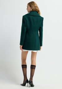 Abrigo de lana verde oscuro, entallado, con un cuello alto y costuras precisas. Combinado con una falda blanca y medias negras hasta la rodilla.